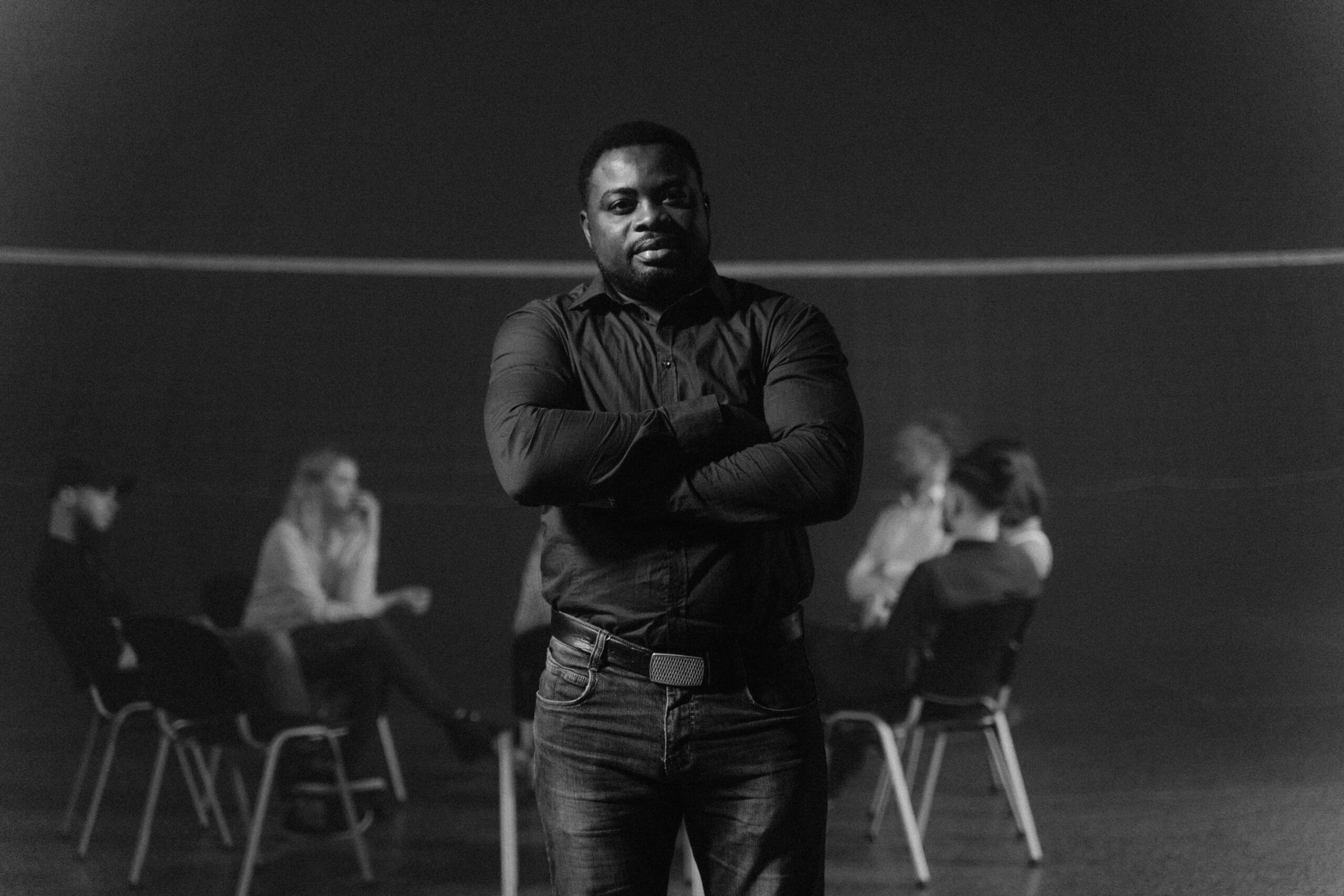 A black and white photo of a man standing with crossed arms in a group meeting.